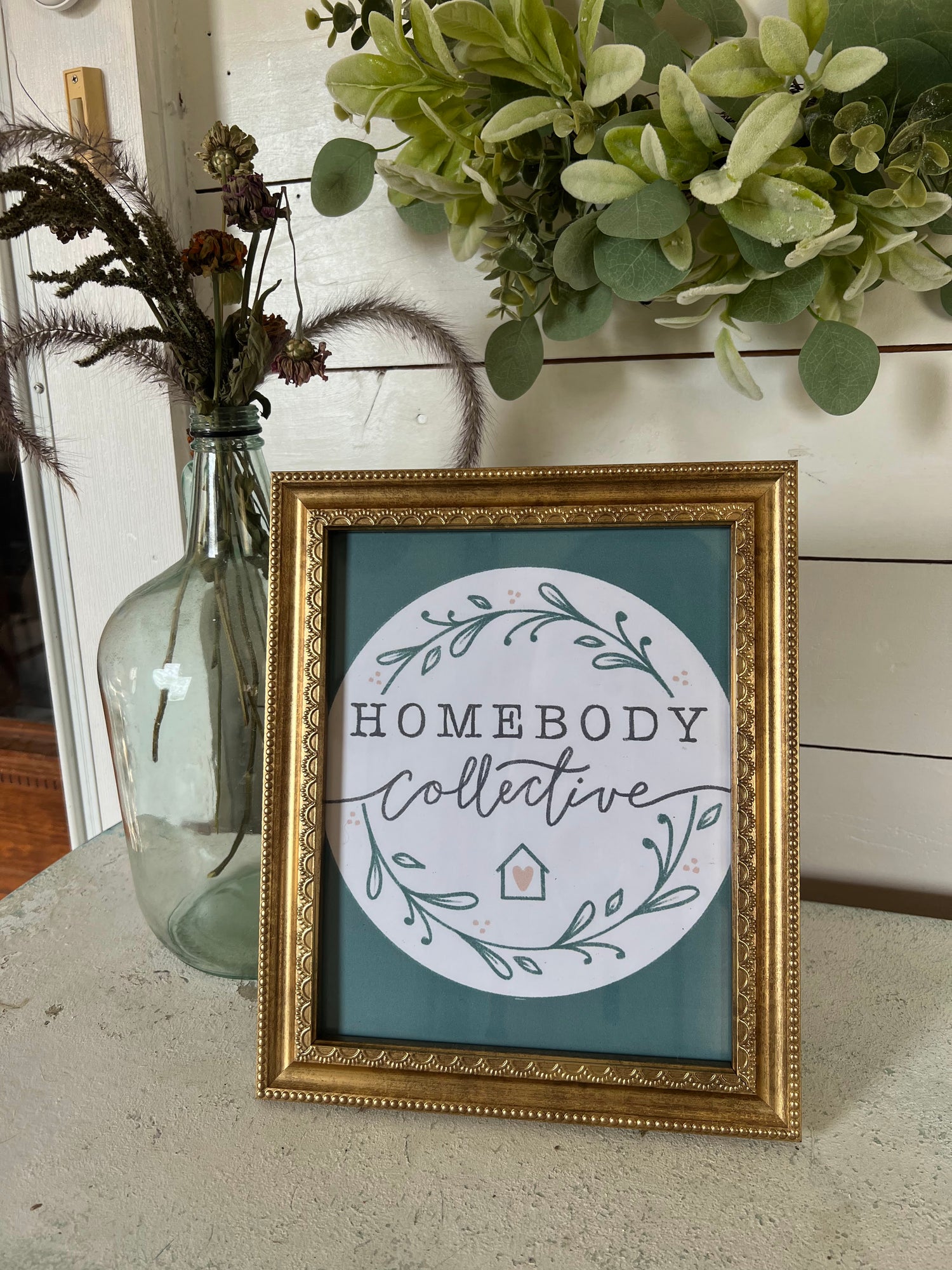 Framed artwork with 'Homebody Collective' on a table next to a plant
